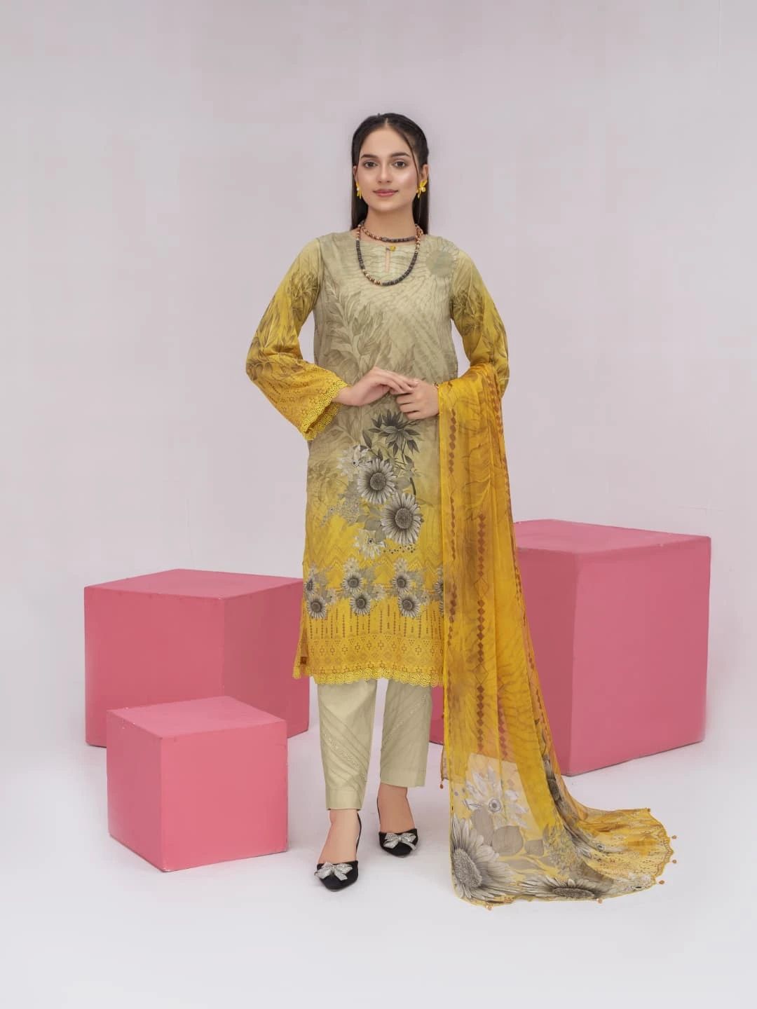 Woman wearing a yellow and green traditional outfit with a floral dupatta against pink cubes on a white background